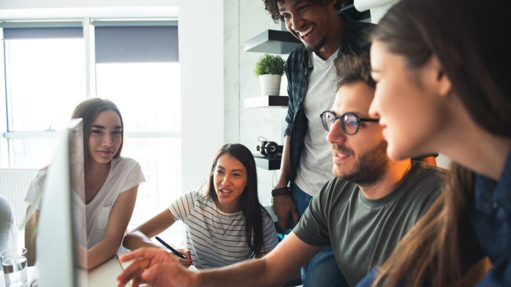 Obrazek przedstawia grupę rozmawiających ludzi, którzy patrzą na coś, co wyświetla się na monitorze komputera