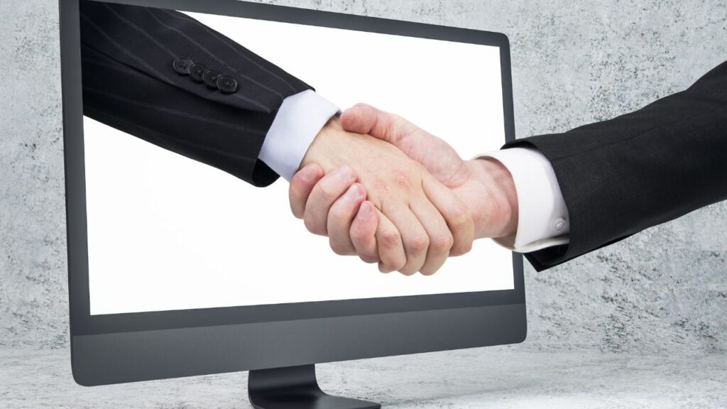 Obrazek przedstawia uścisk dłoni dwóch ludzi. Jedna z nich wychodzi z monitora komputera