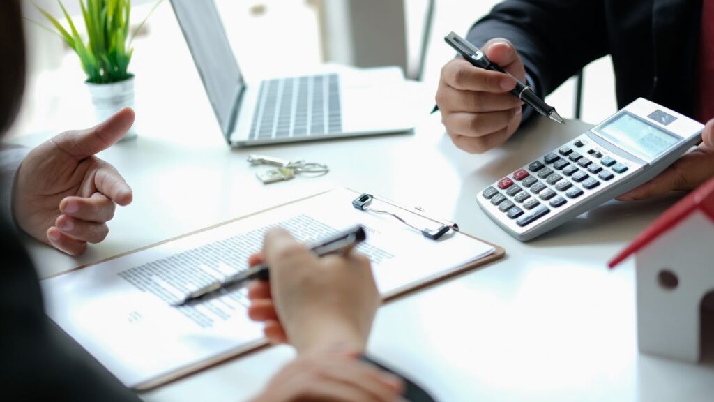 Na obrazku widzimy dłonie dwóch ludzi nad biurkiem. Jedna z osób trzyma długopis gotowa podpisać umowę, druga z nich pokazuje jej wynik na kalkulatorze.